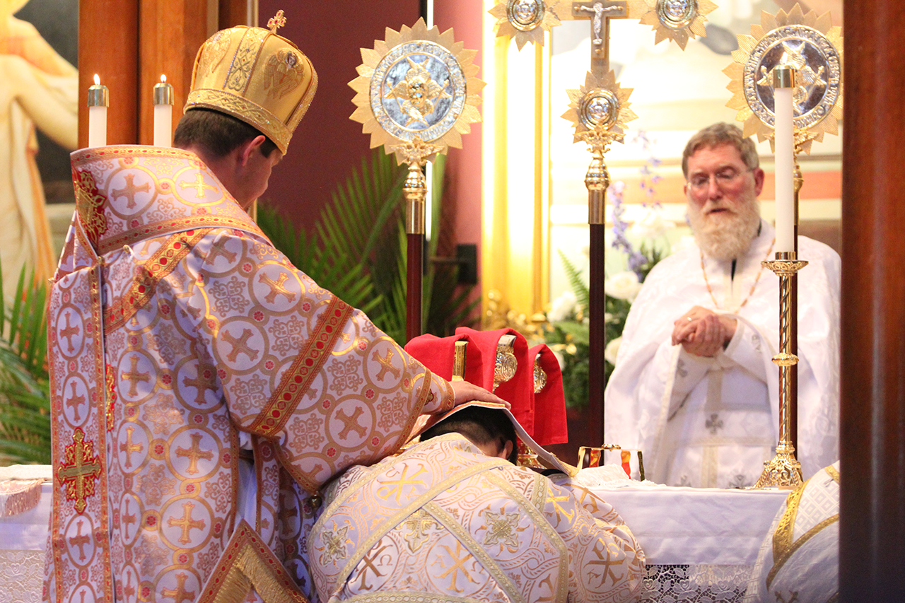 Bishop Bohdan lays his hands on Basilius Magnus' head during the deacon ordination rite, as the deacon candidate kneels and rests his forehead on the altar. Behind the altar, newly-ordained Father Kevin holds the Holy Eucharist in his hands.